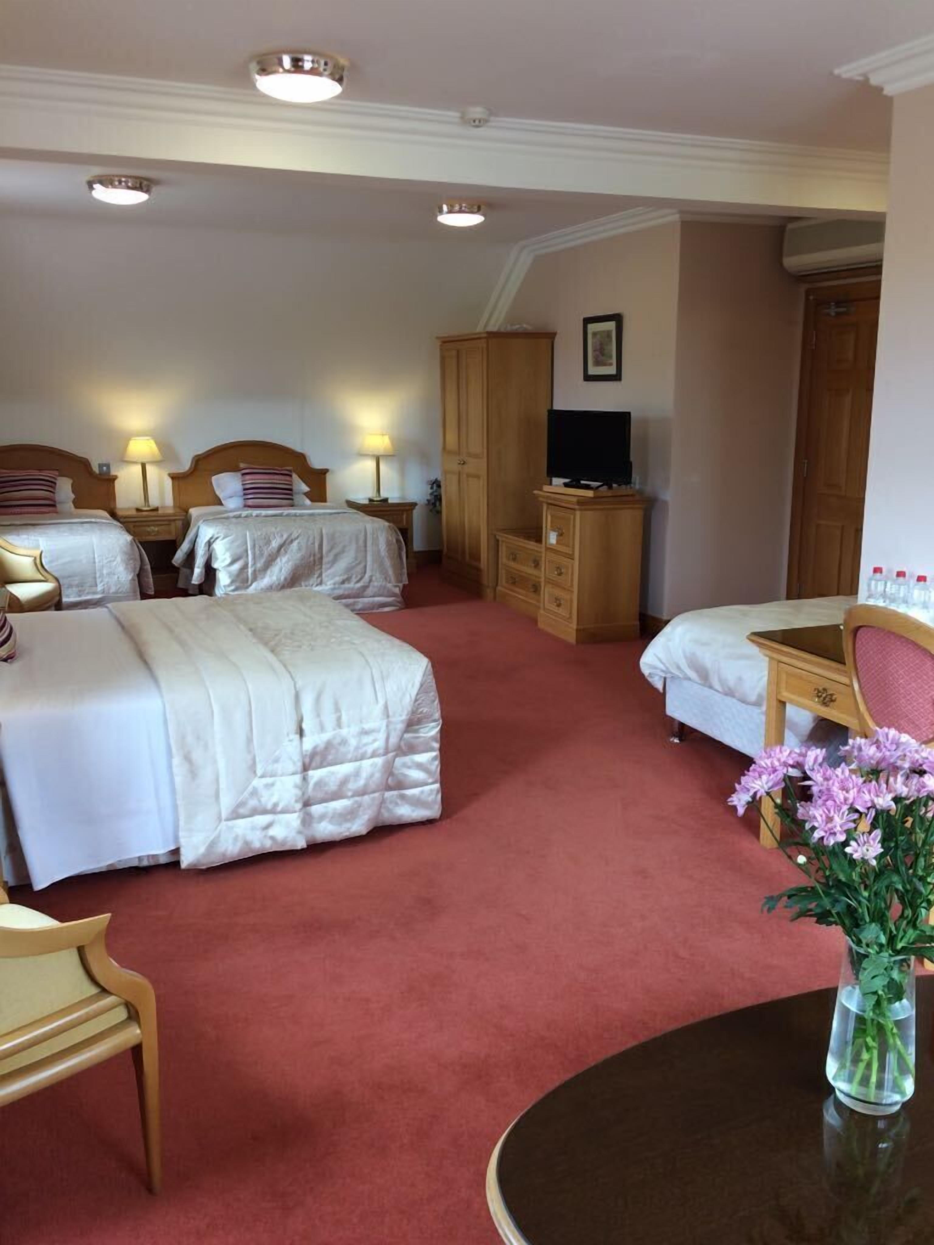 A hotel room with two beds, a desk, a chair, a TV, and a vase with flowers.