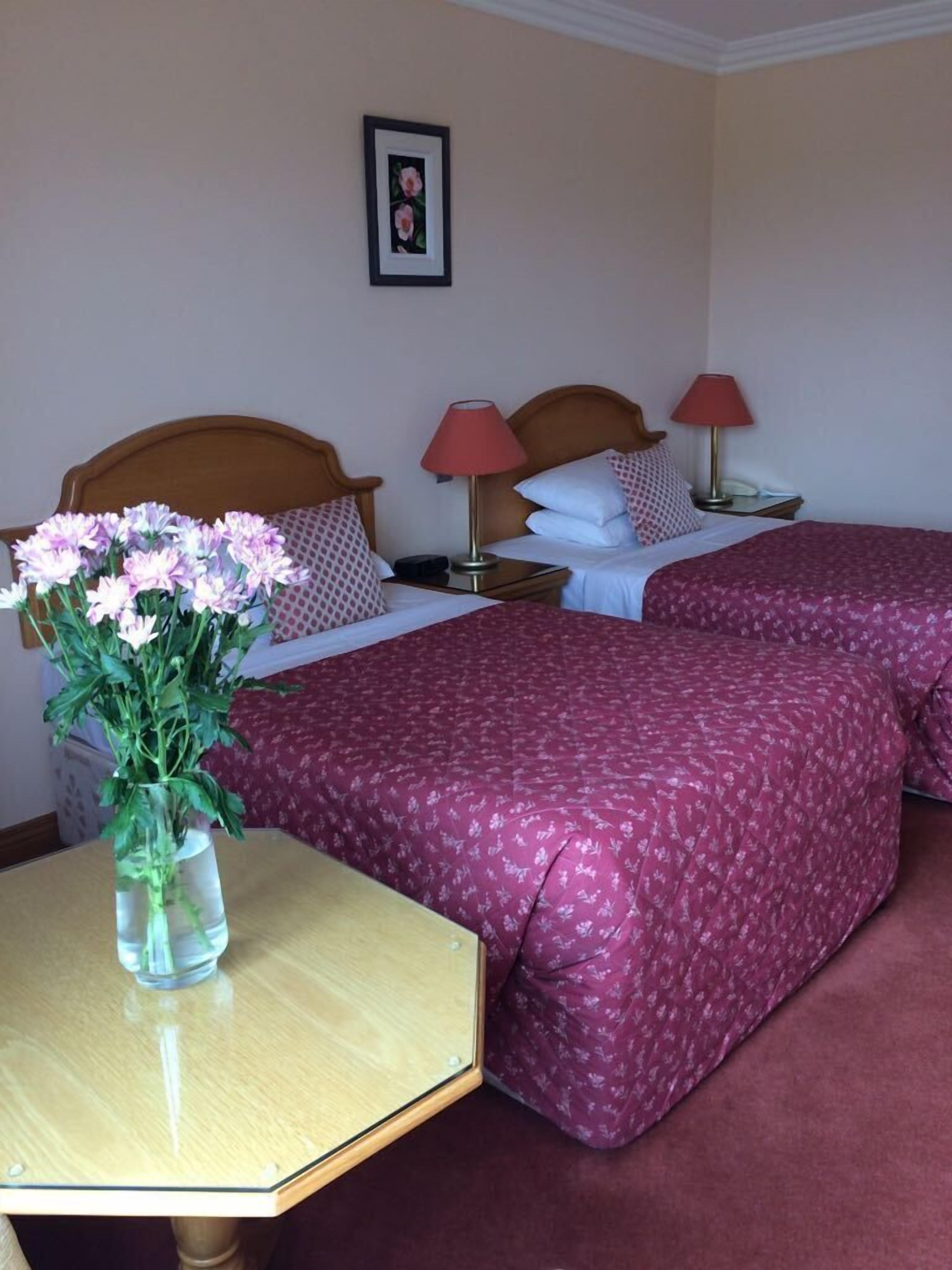 Habitación de hotel con dos camas, cada una con una colcha estampada de flores rojas, cabecero de madera y una mesita de noche con lámpara. Se observa un jarrón con flores sobre una pequeña mesa frente a una de las camas.