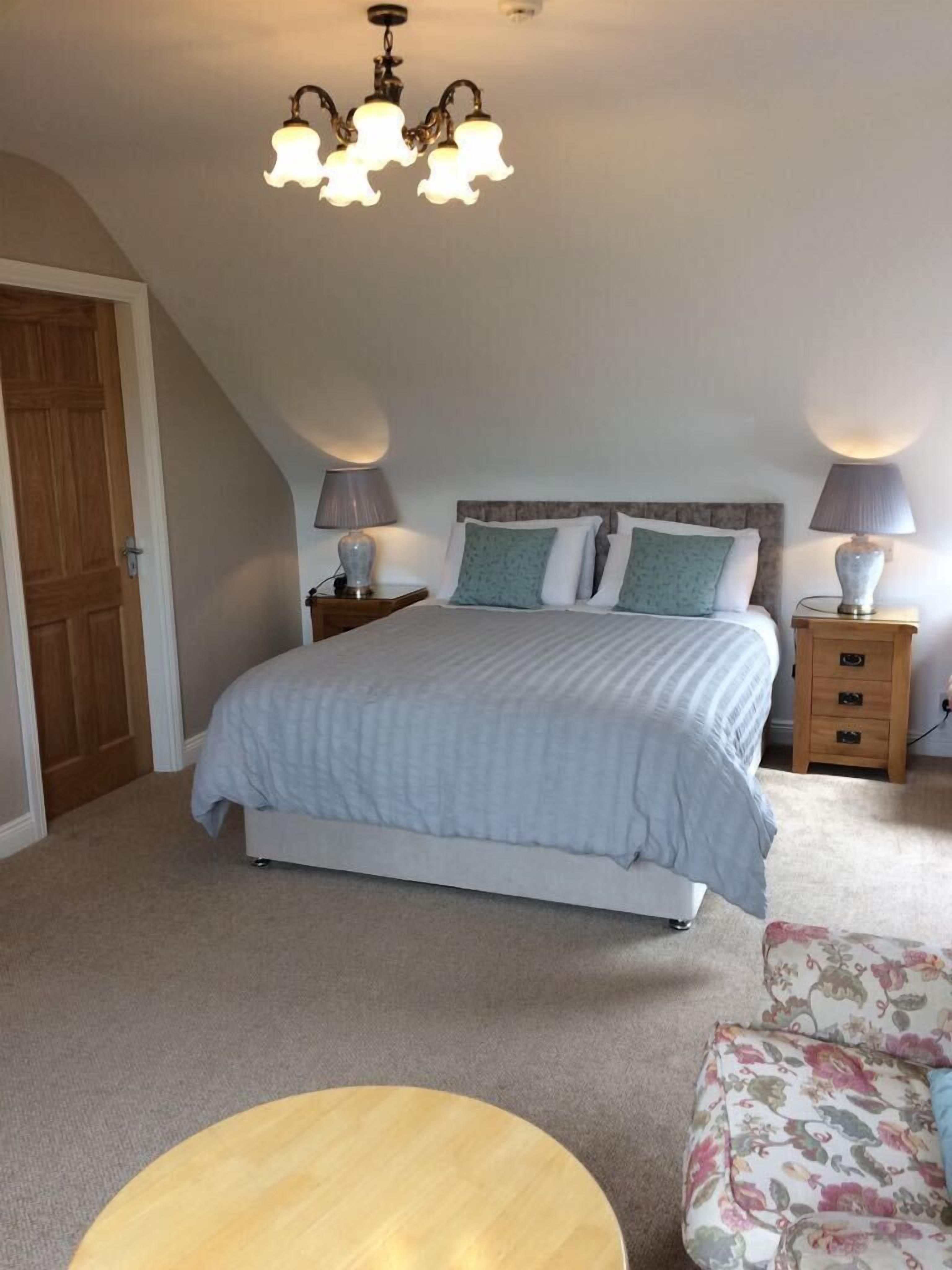 Un dormitorio con una cama, dos mesitas de noche con lámparas, una puerta de madera, una mesa redonda de madera y un sillón con estampado floral.