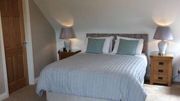 Un dormitorio con una cama, dos mesitas de noche con lámparas, una puerta de madera, una mesa redonda de madera y un sillón con estampado floral.