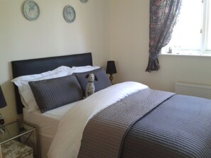 A neatly made bed with a dark headboard, a small table with a lamp beside it, and a window with curtains.