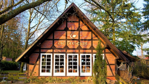 A traditional brick building with a triangular roof, labeled "The Teestube." It features multiple windows and a small garden area with rocks and plants.