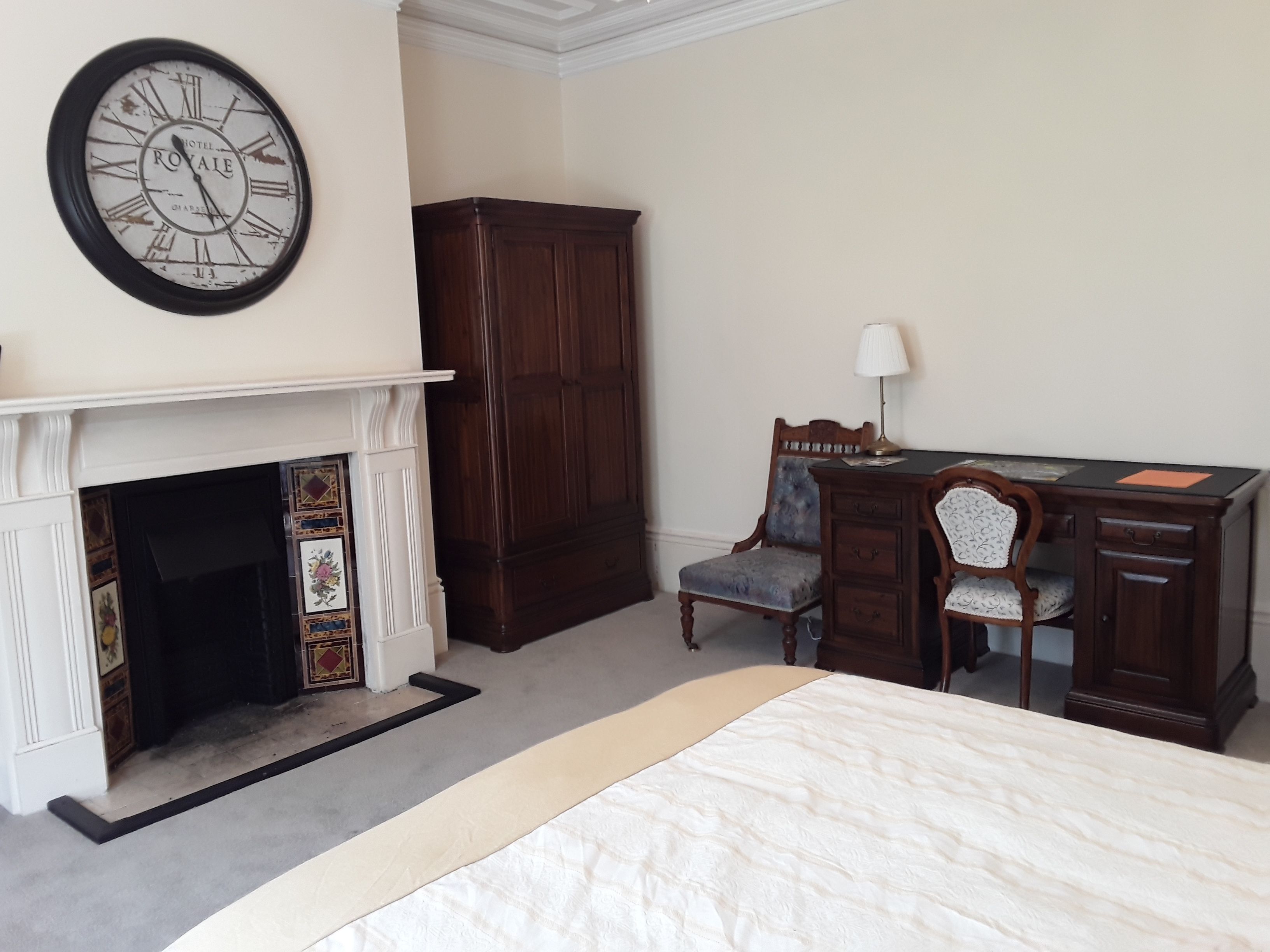 A hotel room with a bed, a desk with a chair, a large wooden wardrobe, and a wall clock.