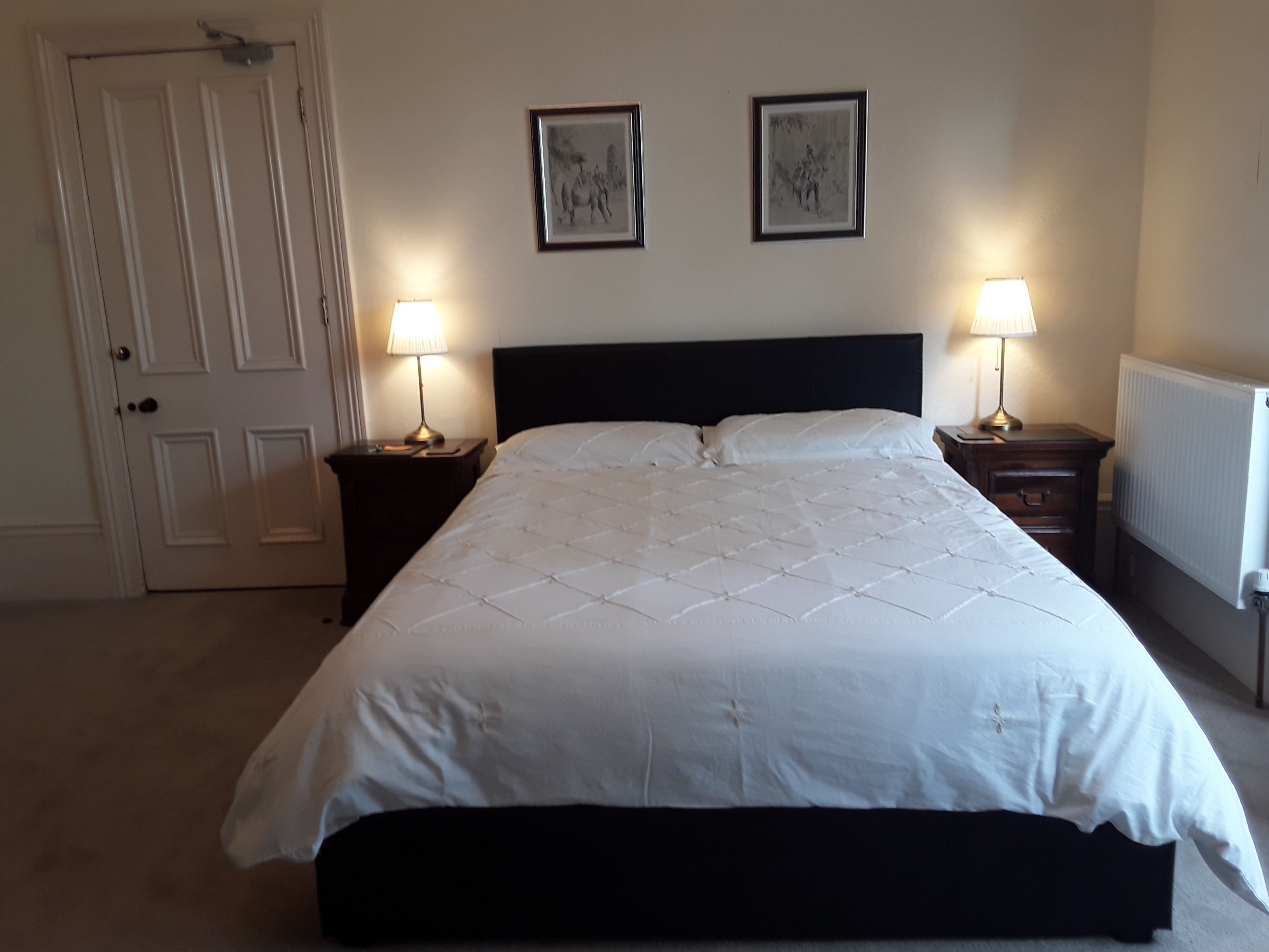 A bedroom with a large bed, two bedside tables with lamps, a door, and two framed pictures on the wall.