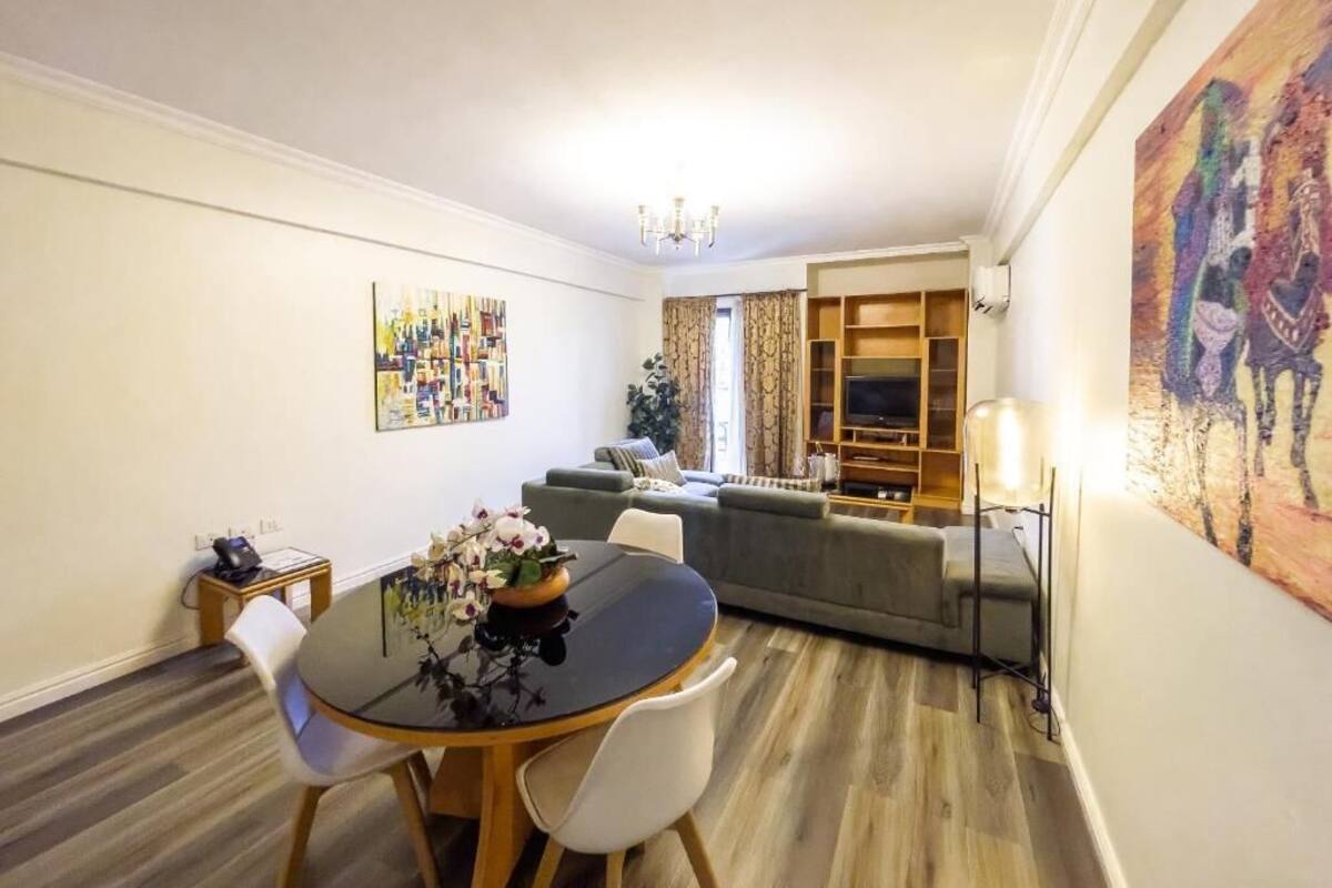 A dining area with a round table, chairs, and a vase of flowers. There is a sofa and a television in the background.