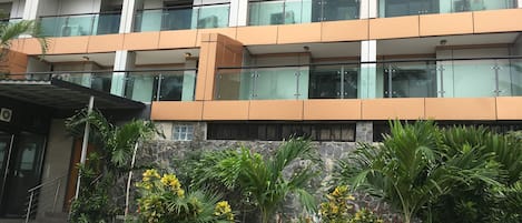 A multi-story building with balconies, a stone facade, and lush greenery in the foreground.