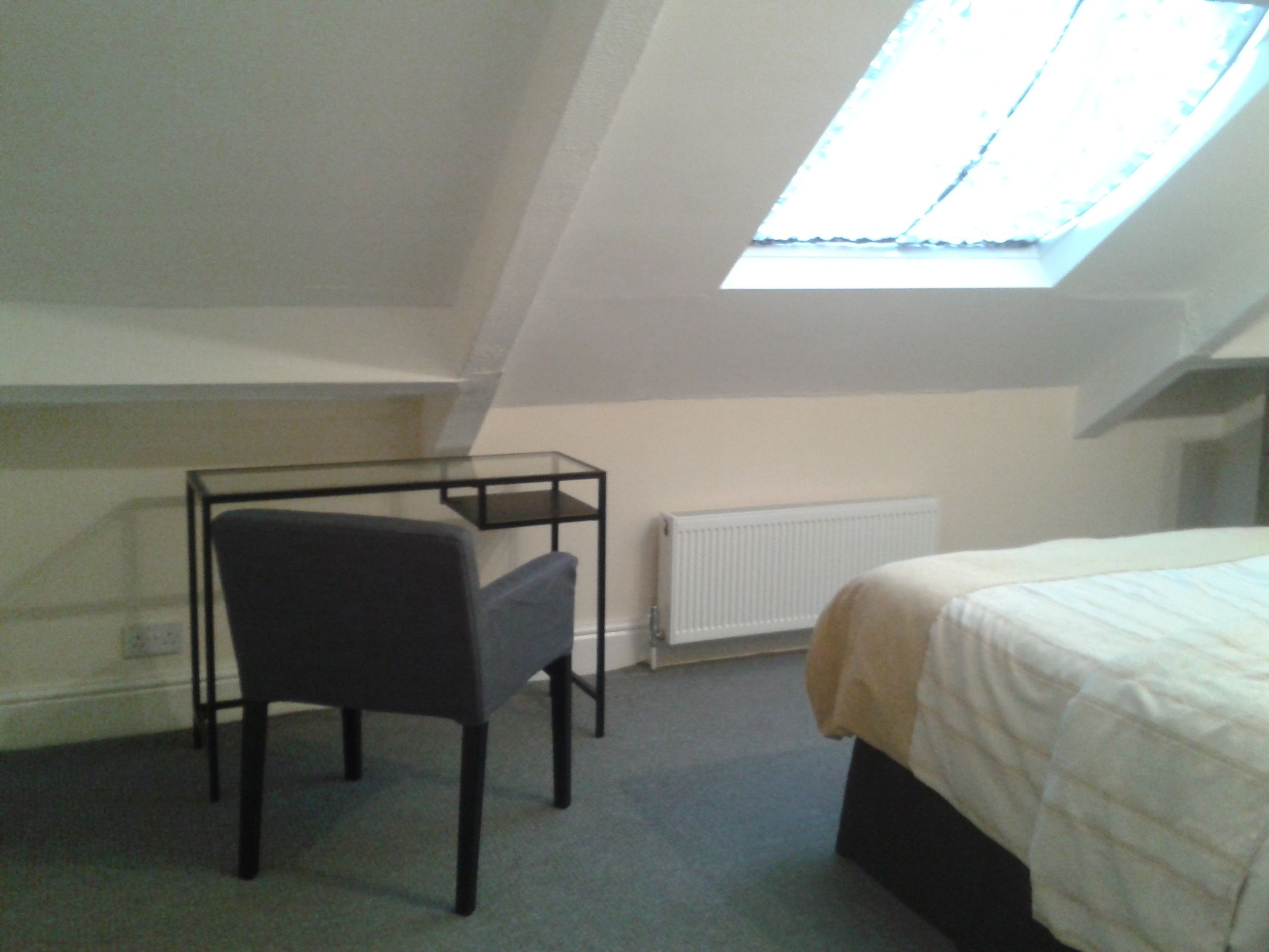 A small, dimly lit room with a single bed, a desk with a chair, and a skylight.