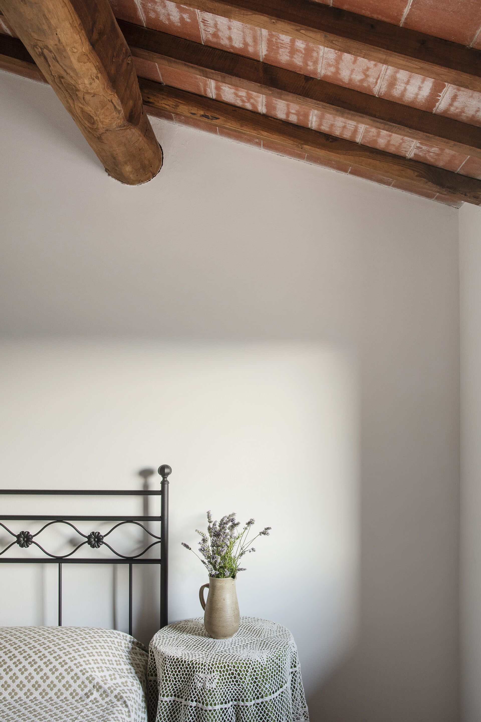 Een slaapkamer met een bed, een klein rond tafeltje met een vaas en een houten plafondbalk.
