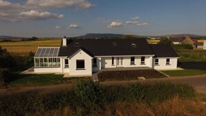 Une maison moderne à deux étages, dotée d’une grande véranda en verre, entourée de vastes champs à perte de vue et surplombant un ciel dégagé.