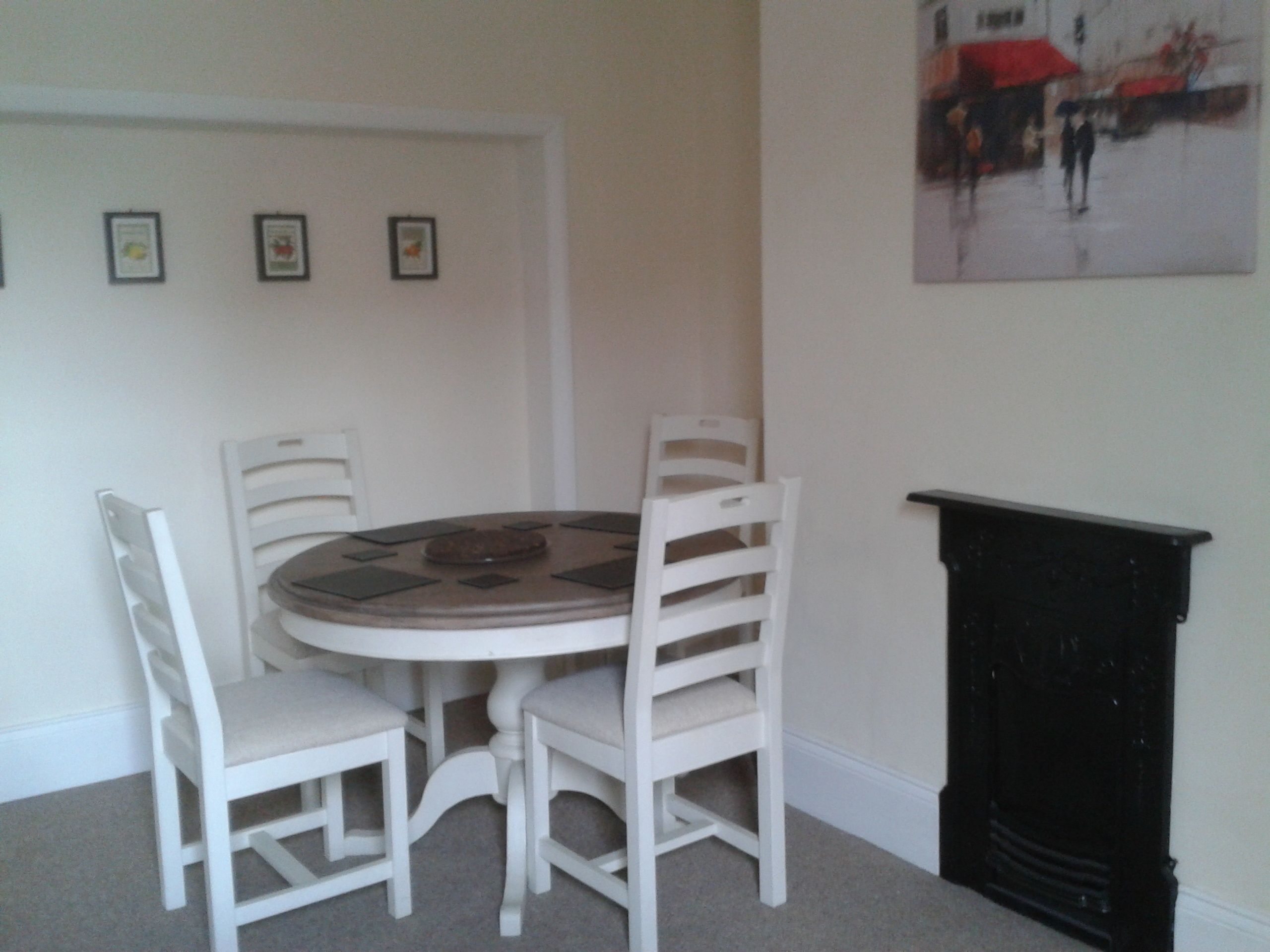 Een eethoek met een ronde tafel en vier witte stoelen. Rechts bevindt zich een zwarte schoorsteen en boven de tafel hangt een lijst met een afbeelding.