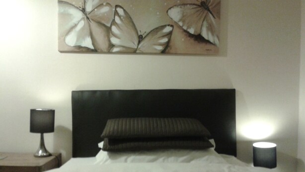 Una cama bien hecha con un cabecero oscuro, dos almohadas y una manta blanca. Encima de la cama, un cuadro con mariposas. Dos lámparas de noche encendidas, una a cada lado de la cama.