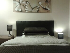 A neatly made bed with a dark headboard, two pillows, and a white blanket. There is a wall-mounted artwork of butterflies above the bed. Two bedside lamps are on, one on each side of the bed. - Park View (Gateshead)