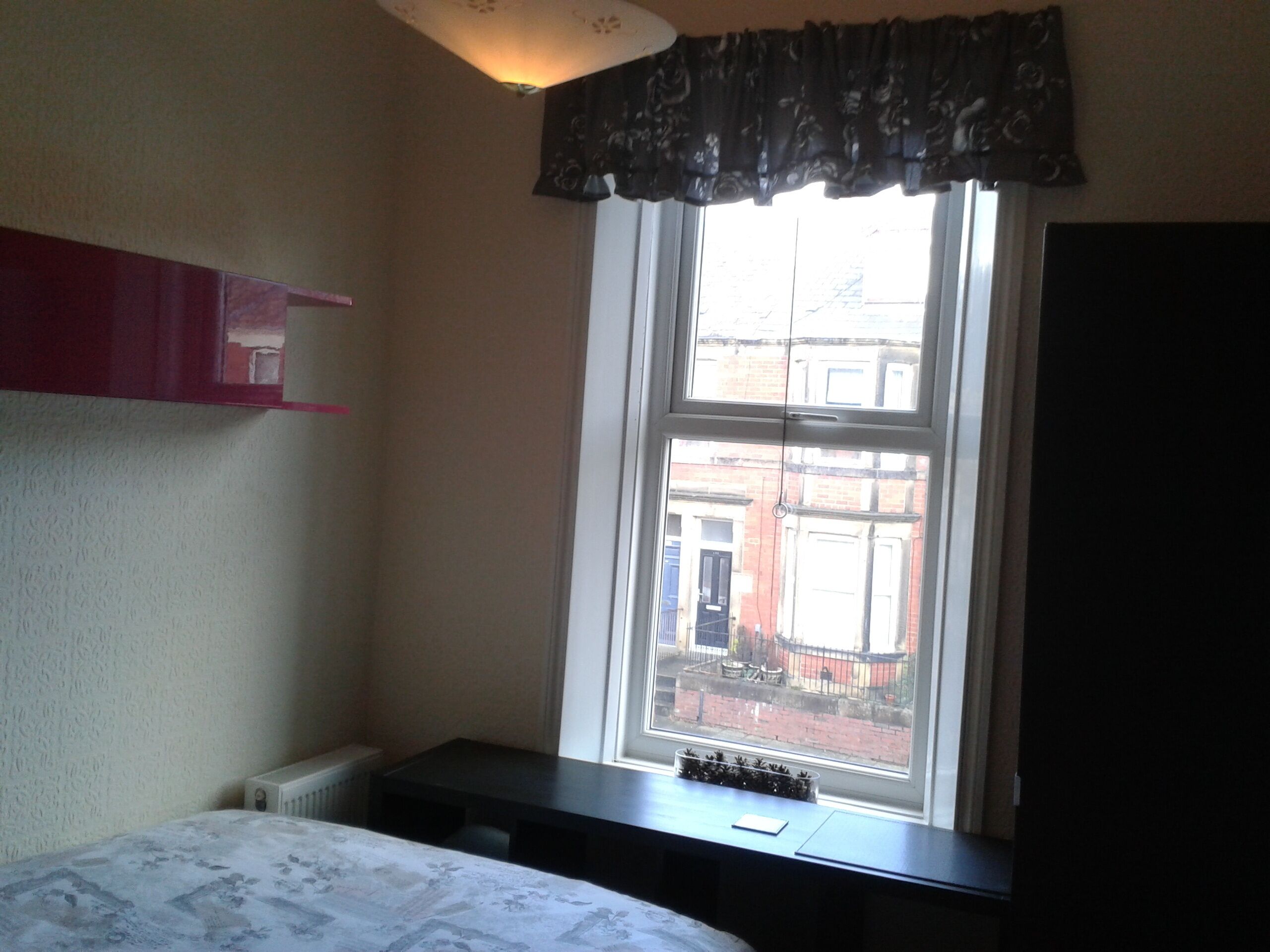 Une chambre à coucher comprenant un lit, un bureau, un radiateur, une fenêtre donnant sur des immeubles et un luminaire au plafond.