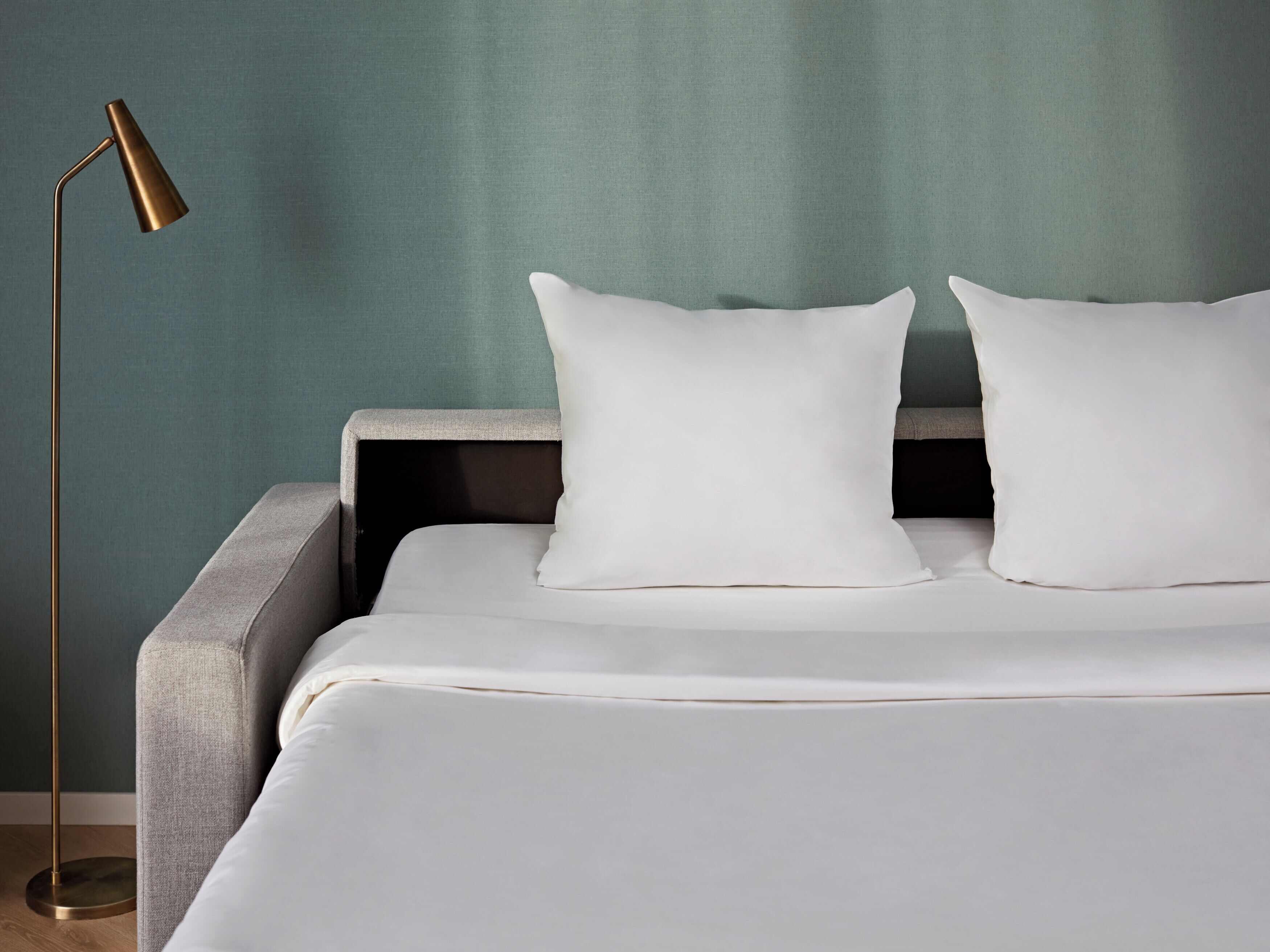 A neatly made bed with white linens, a grey headboard, and two white pillows. A modern floor lamp with a gold base is positioned to the left of the bed.
