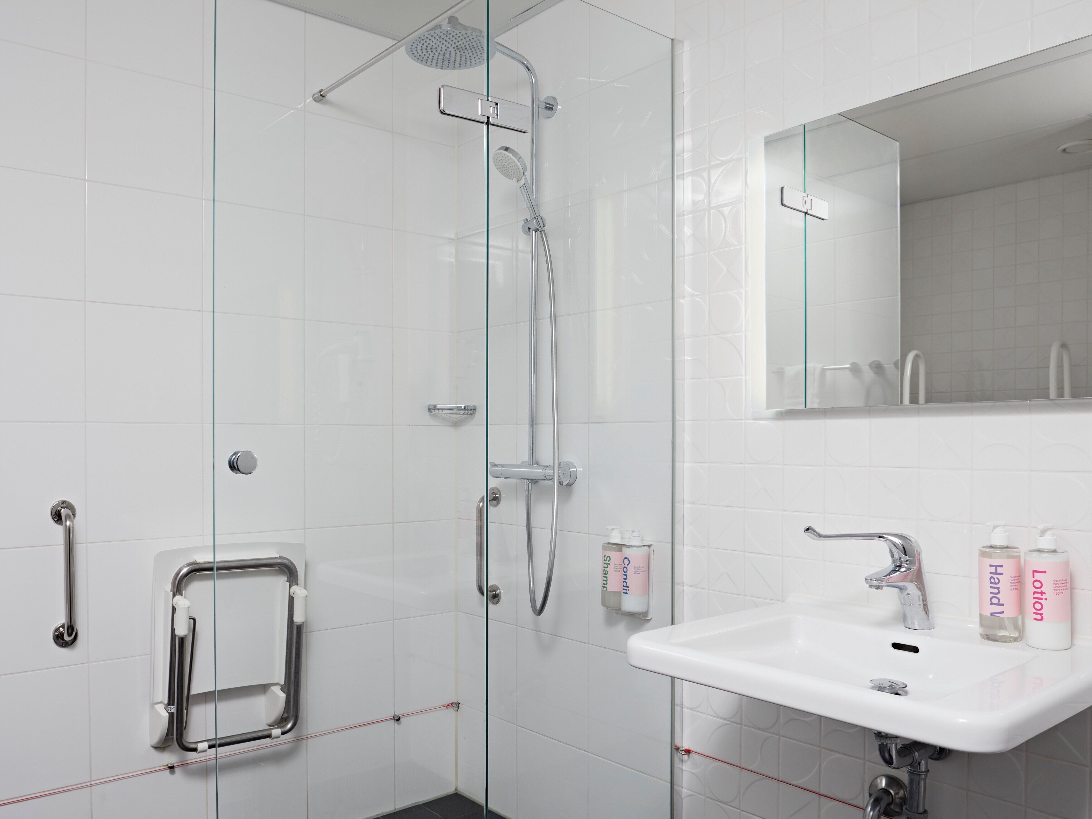 Une salle de bain moderne avec une douche à l’italienne en verre, un lavabo blanc avec un miroir au-dessus, et des articles de toilette posés sur le lavabo.
