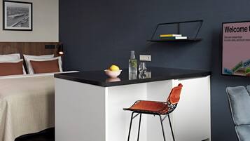 A modern hotel room with a bed, a small kitchen area, a bar stool, and a framed welcome sign.