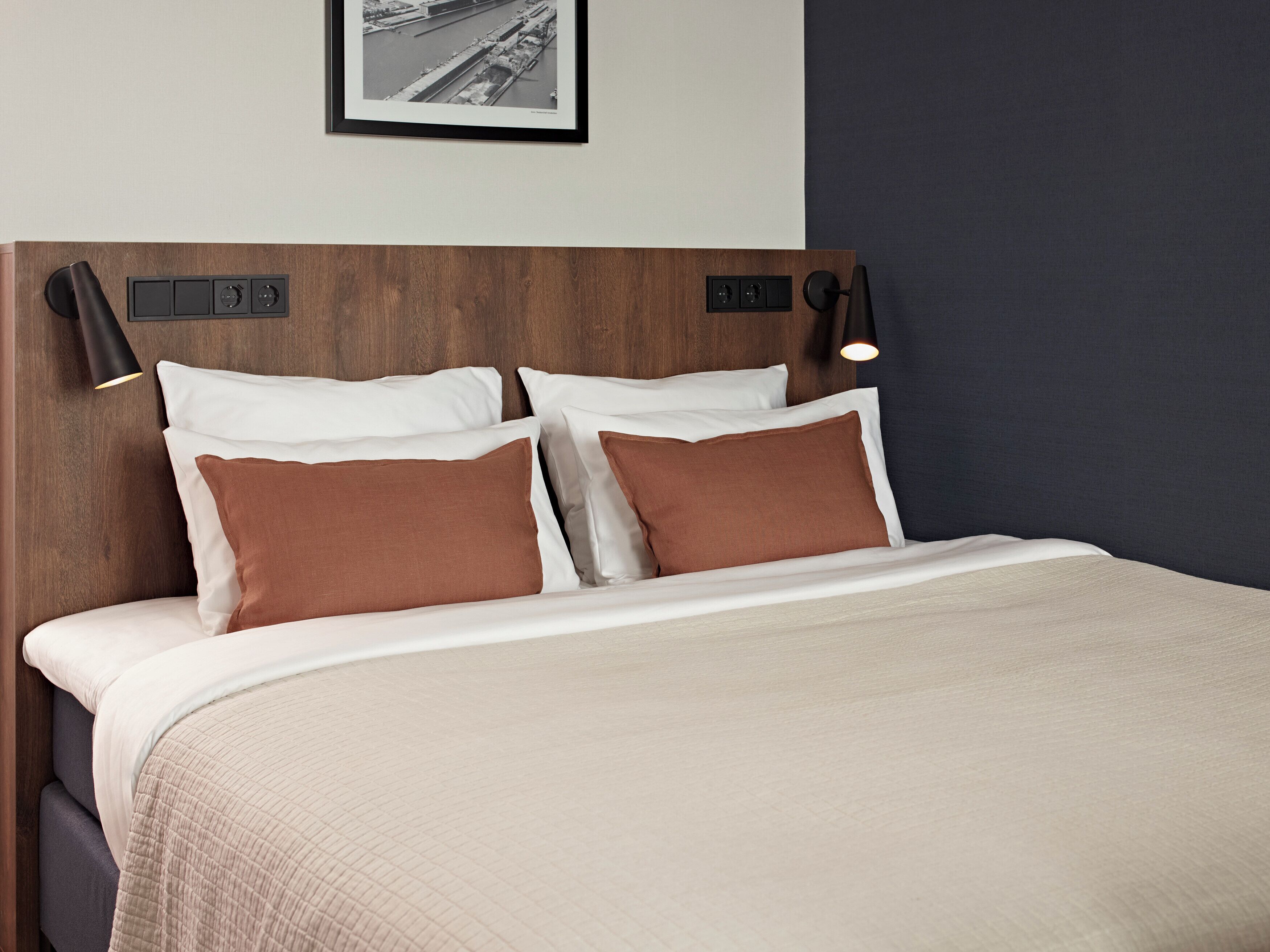 A neatly made bed with a beige quilt, two white pillows, and two brown pillows. There are two black wall-mounted lamps on either side of the bed.