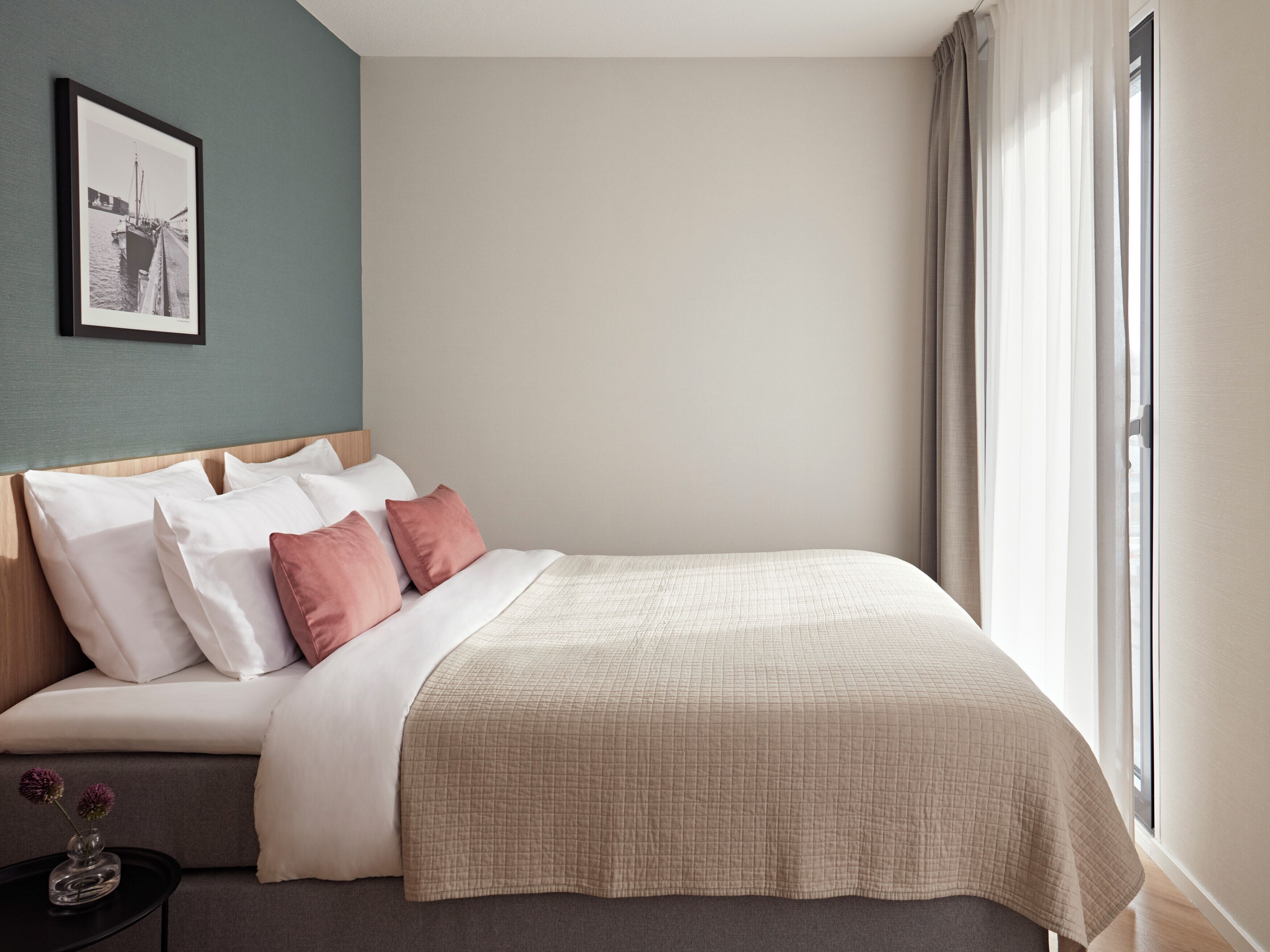 A neatly made bed with white and pink pillows, a beige quilted blanket, and a small table with a vase of flowers.