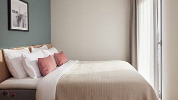A neatly made bed with white and pink pillows, a beige quilted blanket, and a small table with a vase of flowers.