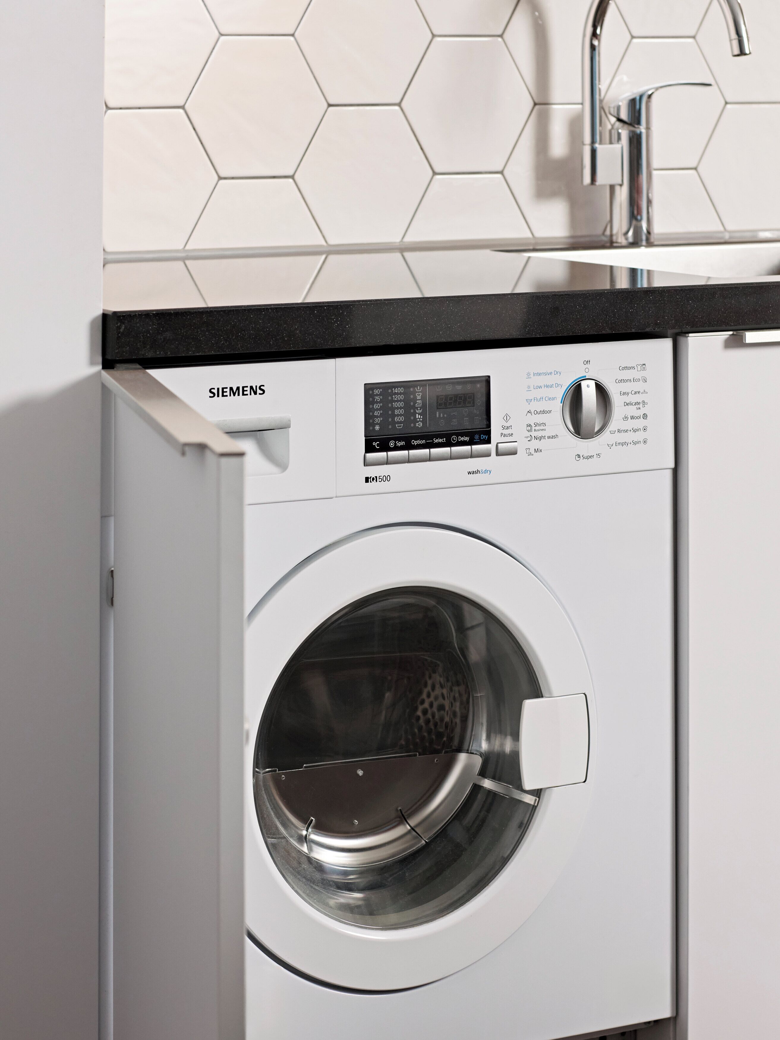 A front-loading Siemens washing machine with a digital control panel, set in a kitchen with a hexagonal tile backsplash and a black countertop.