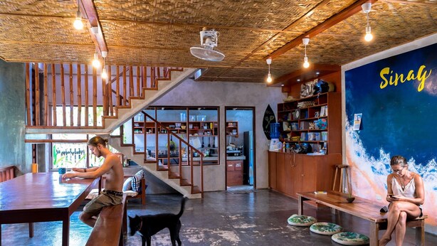 A spacious room with a wooden ceiling, a staircase, a large wooden table, and a wall with a 'Sinag' sign. - Sinag Hostel - Adults Only (General Luna)