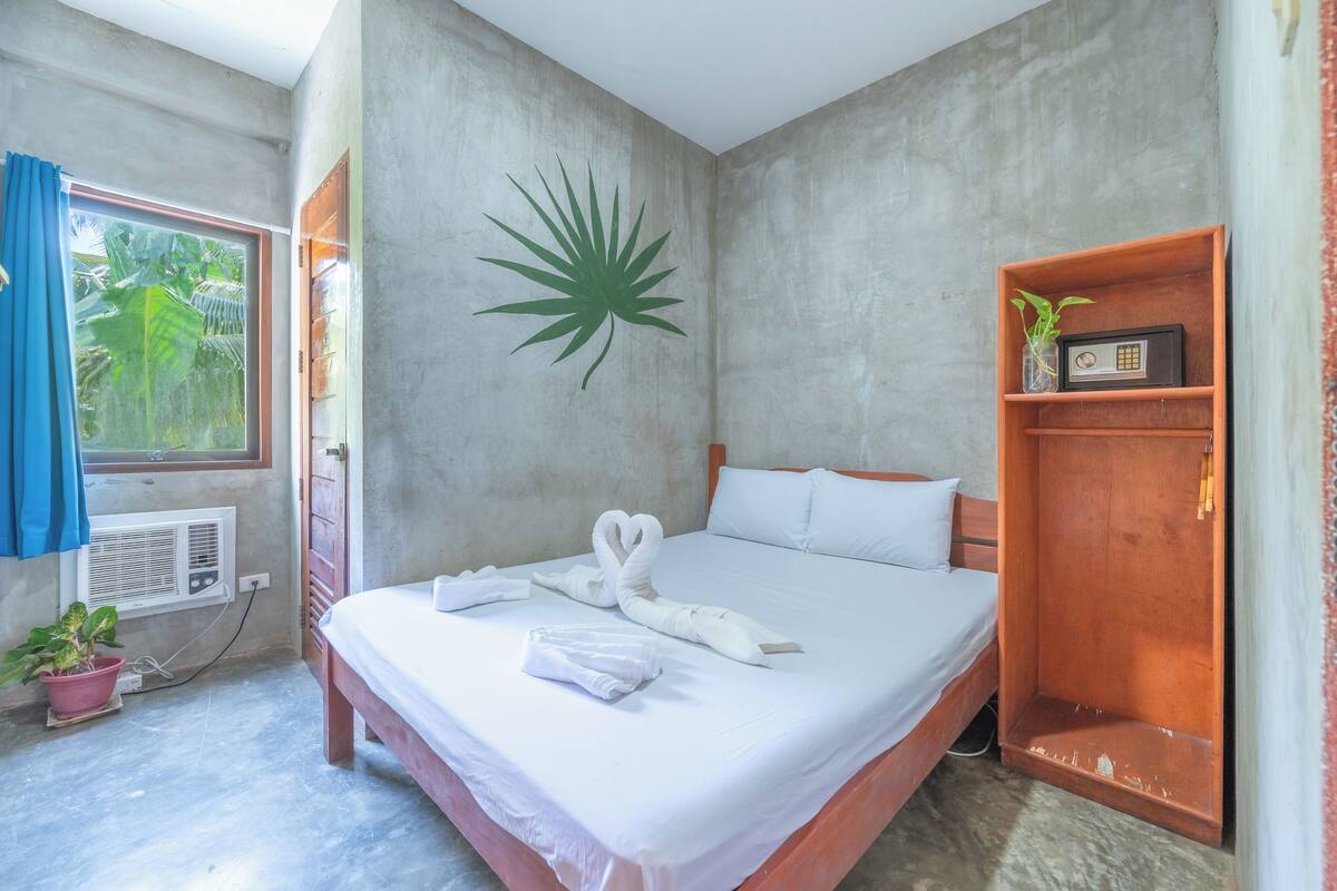 A hotel room with a bed, a wall-mounted plant, a wooden shelf with a radio, and a window with curtains.