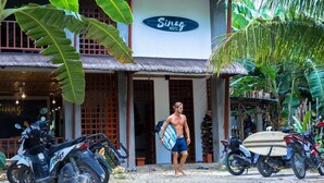A man carrying a surfboard exiting a building labeled "Sinag Hostel." - Sinag Hostel - Adults Only (General Luna)