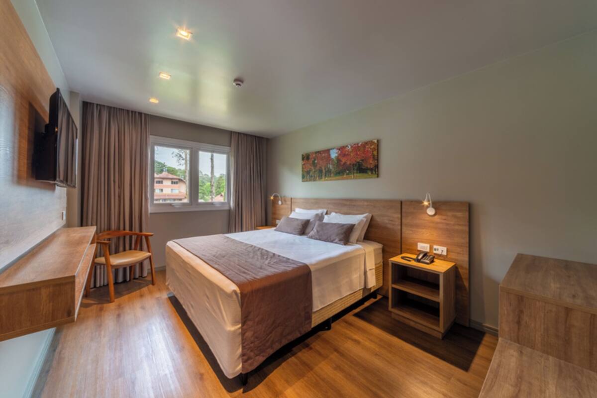 A modern hotel room with a large bed, a desk, a chair, a bedside table with a lamp, and a painting on the wall.