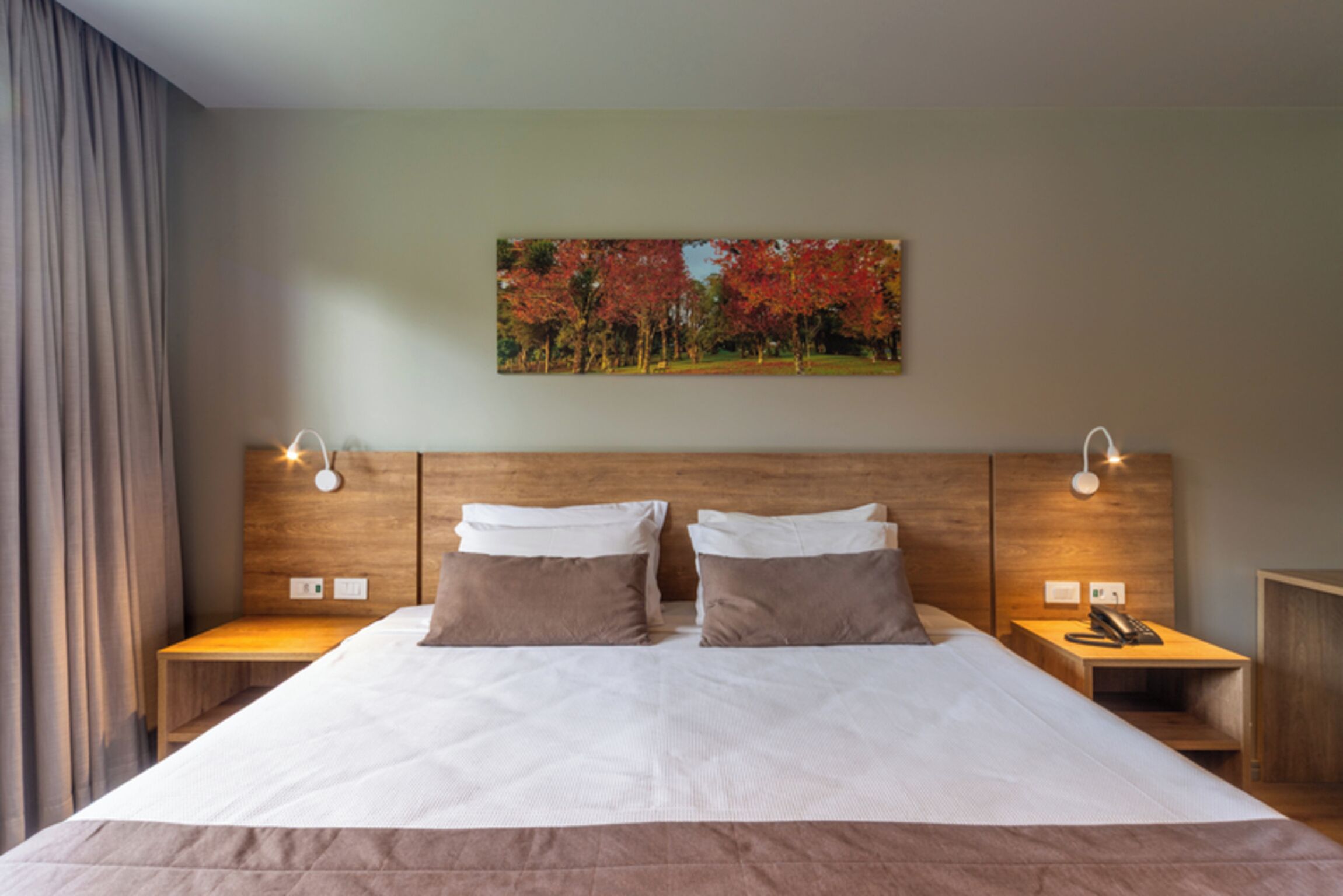 Una cama bien hecha con dos almohadas, un cabecero con iluminación integrada y un cuadro de árboles otoñales en la pared.