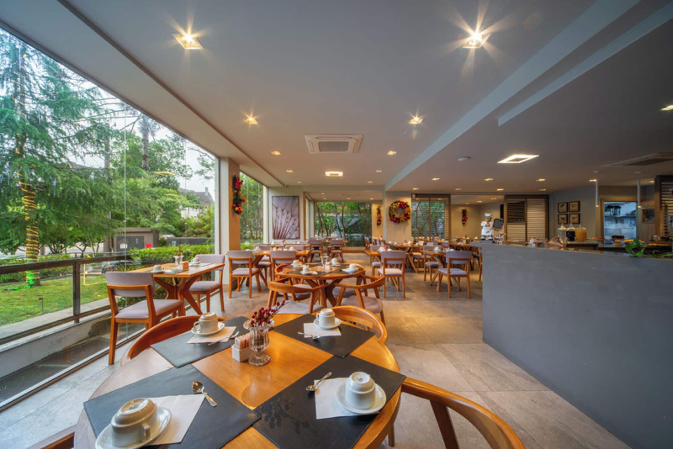 A spacious indoor dining area with multiple tables and chairs, large windows offering a view of the outdoors, and a modern ceiling design with recessed lighting.