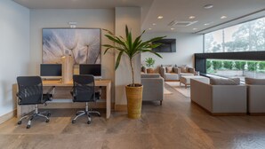 A modern office space with two desks, black office chairs, a large potted plant, a sofa, and a television on the wall. - Hotel Laghetto Fratello (Gramado)