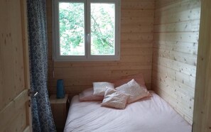Une petite pièce chaleureuse, avec un intérieur en bois, un lit recouvert d’une literie rose, une fenêtre donnant sur la verdure et un rideau à motifs.