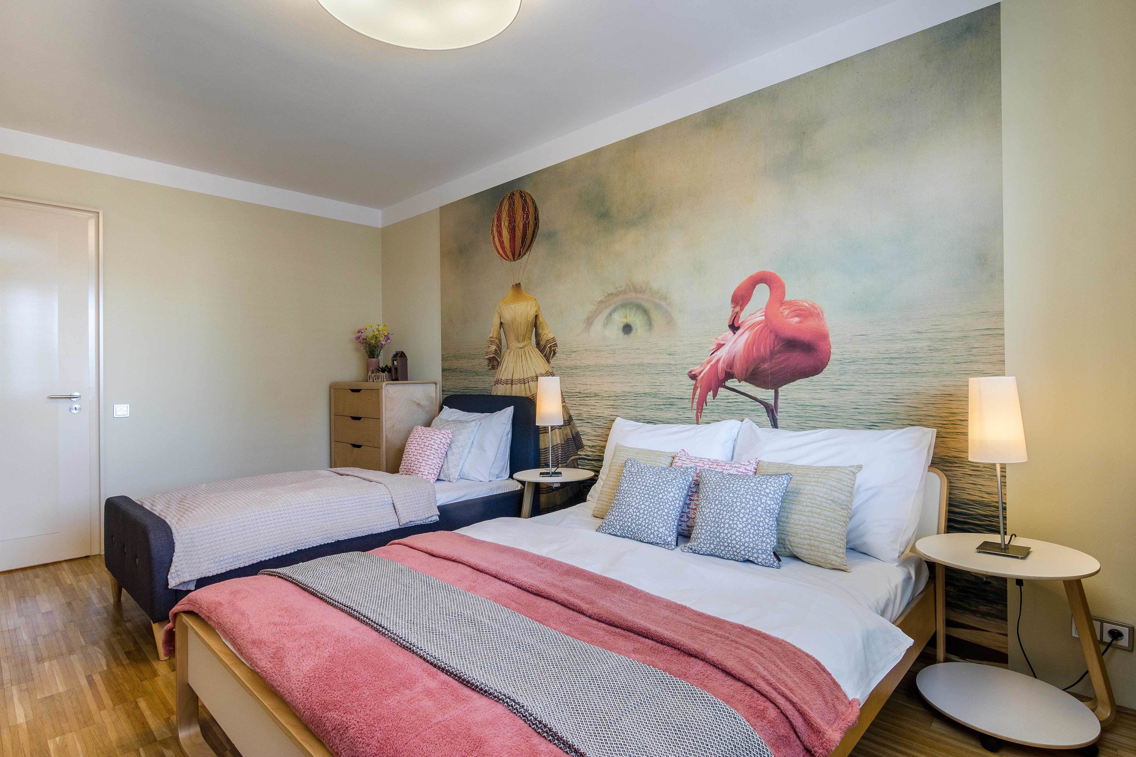 A hotel room with two beds, a mural of a flamingo and an eye, and a small round table with a lamp.