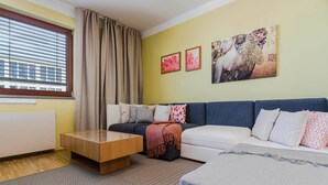 Un salon moderne avec un canapé, une table basse et un lit. Il y a deux tableaux au mur, une fenêtre avec des rideaux et un radiateur.