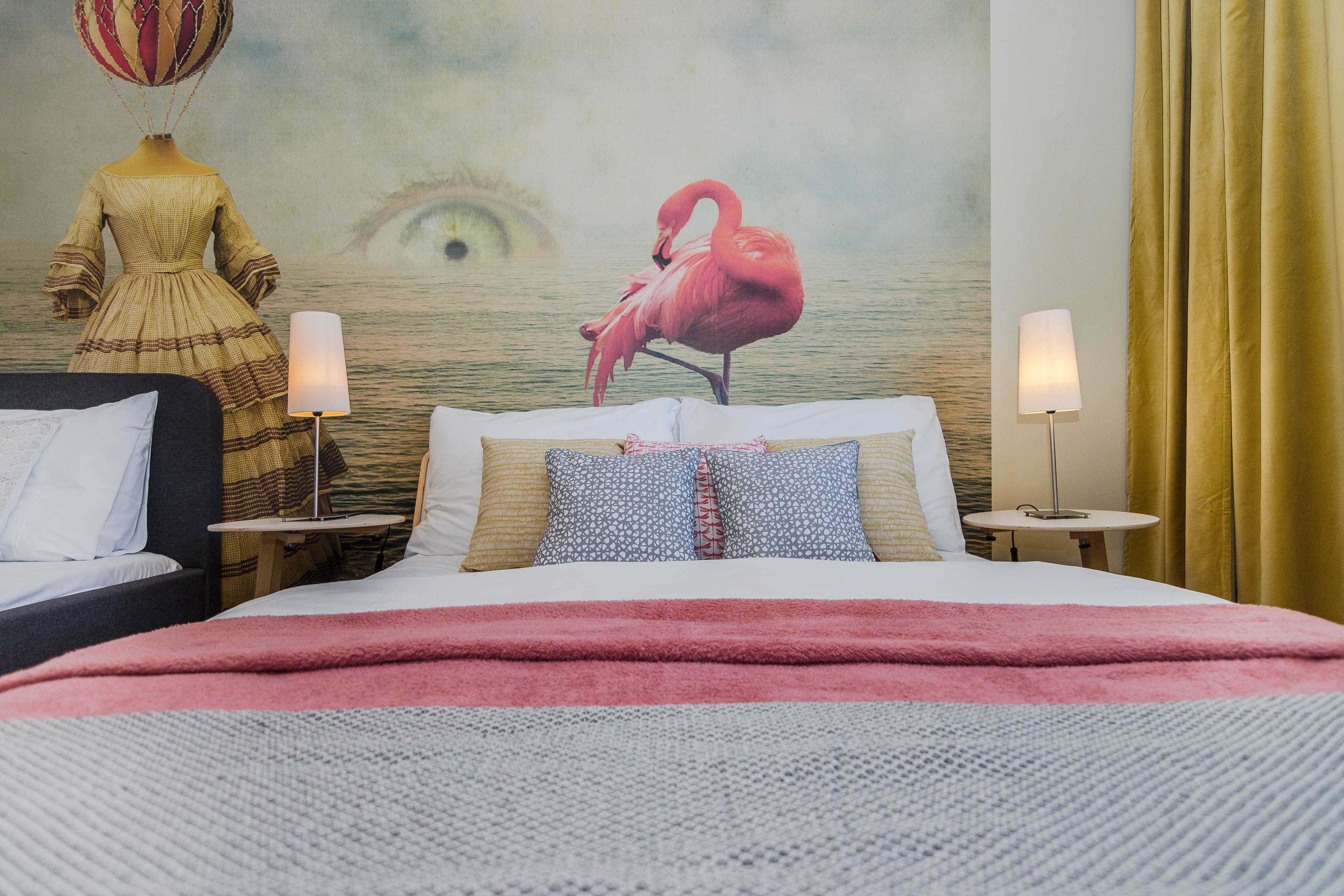 Habitación de hotel con una cama grande, una mesita de noche con lámpara, un mural en la pared que muestra un flamenco y un ojo, y una ventana con cortinas.