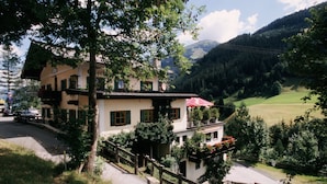 Un bâtiment de style chalet traditionnel, doté d’un balcon et entouré d’une végétation luxuriante, avec une chaîne de montagnes en toile de fond.