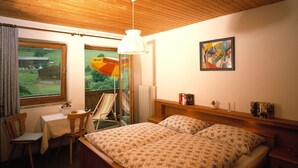 Une chambre avec un plafond en bois, un lit recouvert d’une literie à motifs floraux, une chaise, une table, une fenêtre donnant sur une grange et un tableau accroché au mur.