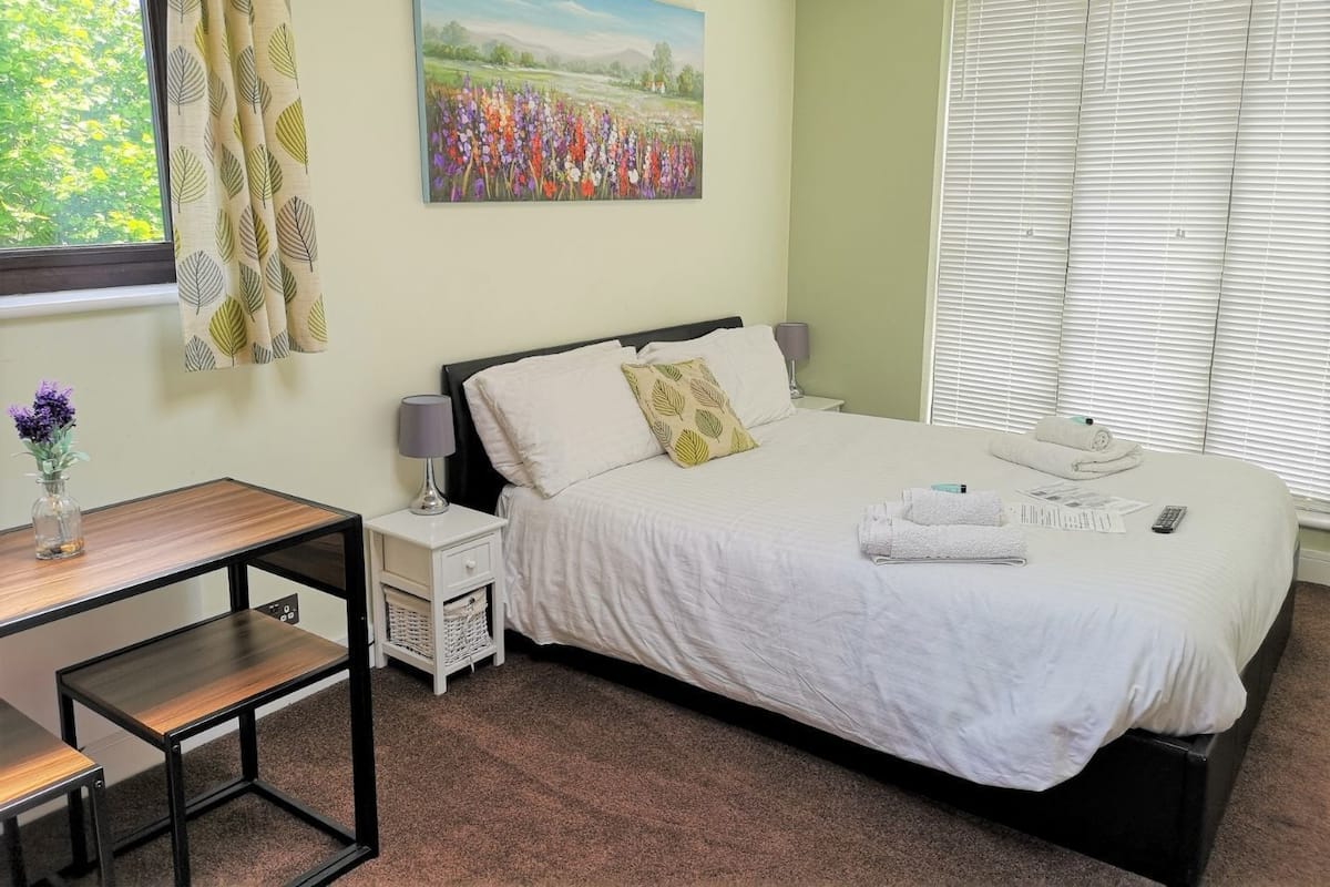 Quarto com cama, mesas de cabeceira, escrivaninha e janela com cortinas.