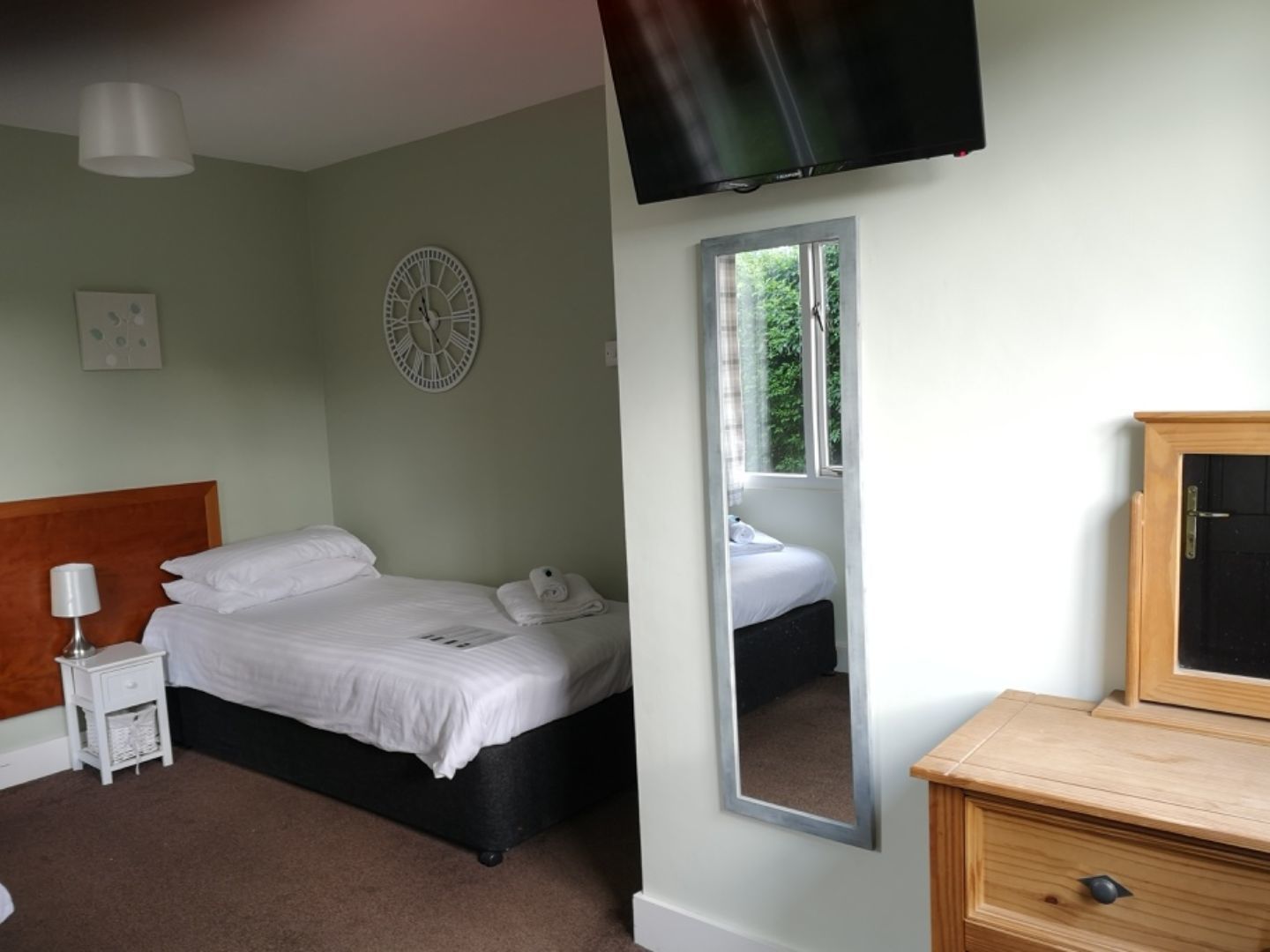 A bedroom with a bed, a bedside table with a lamp, a wall clock, a mirror, and a TV mounted on the wall.
