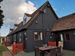 A traditional building with a dark exterior, a sloped roof, and a visible chimney. There is a wooden fence in front, and a table with chairs outside. - Swedish Cottages (Luton)