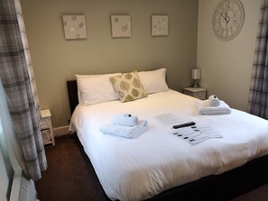 A neatly made bed with white linens, a pillow with a leaf pattern, and two rolled towels. There is a clock on the wall, a remote control, and a menu on the bed. - Swedish Cottages (Luton)
