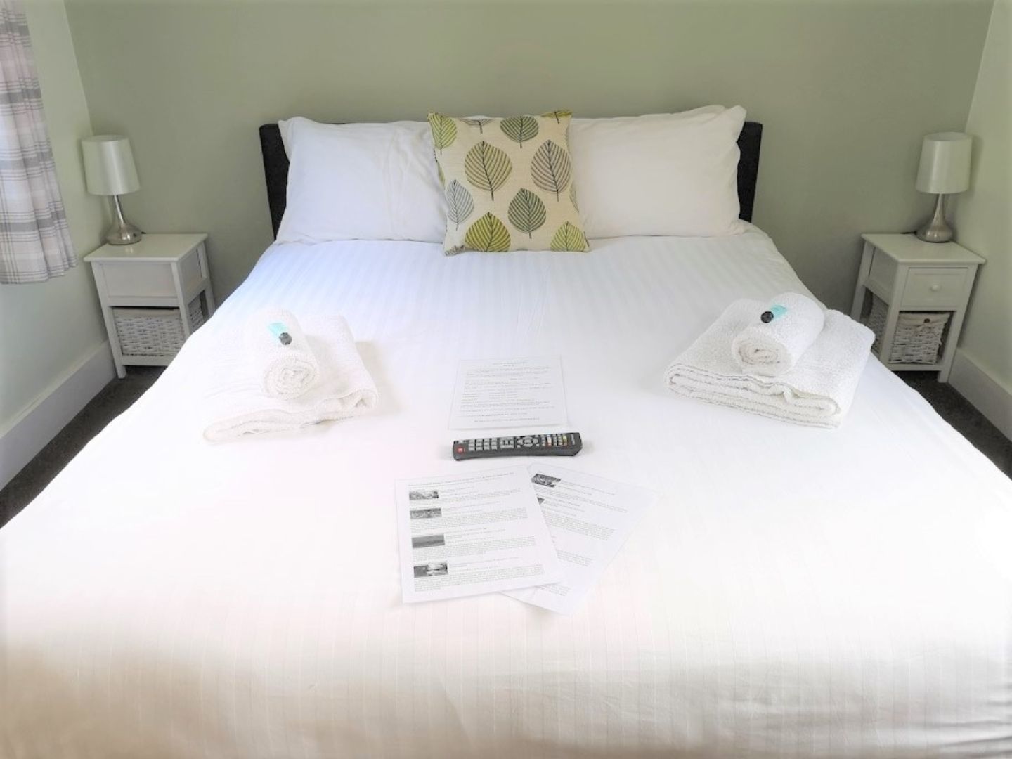 A neatly made bed with white linens, two pillows, a remote control, and a set of papers on the bed.