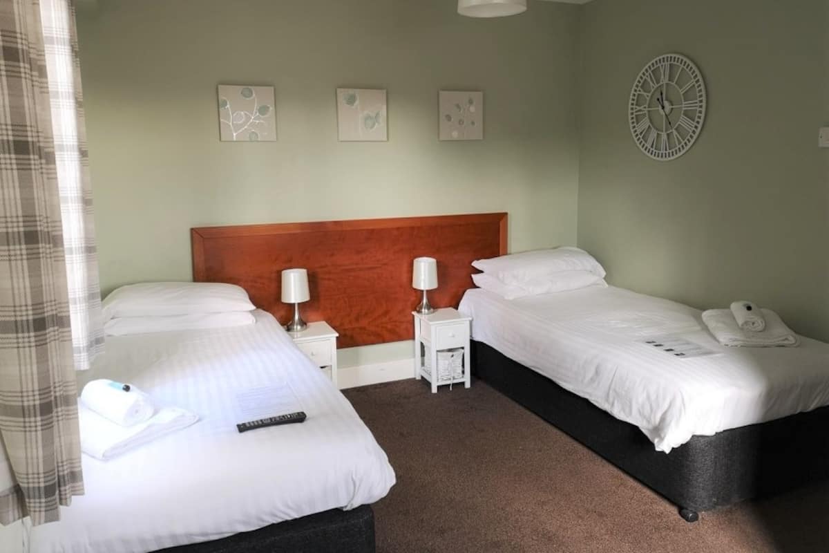 Quarto de hotel com duas camas, um painel de cabeceira de madeira, um relógio na parede e uma pequena mesa de cabeceira com abajur.