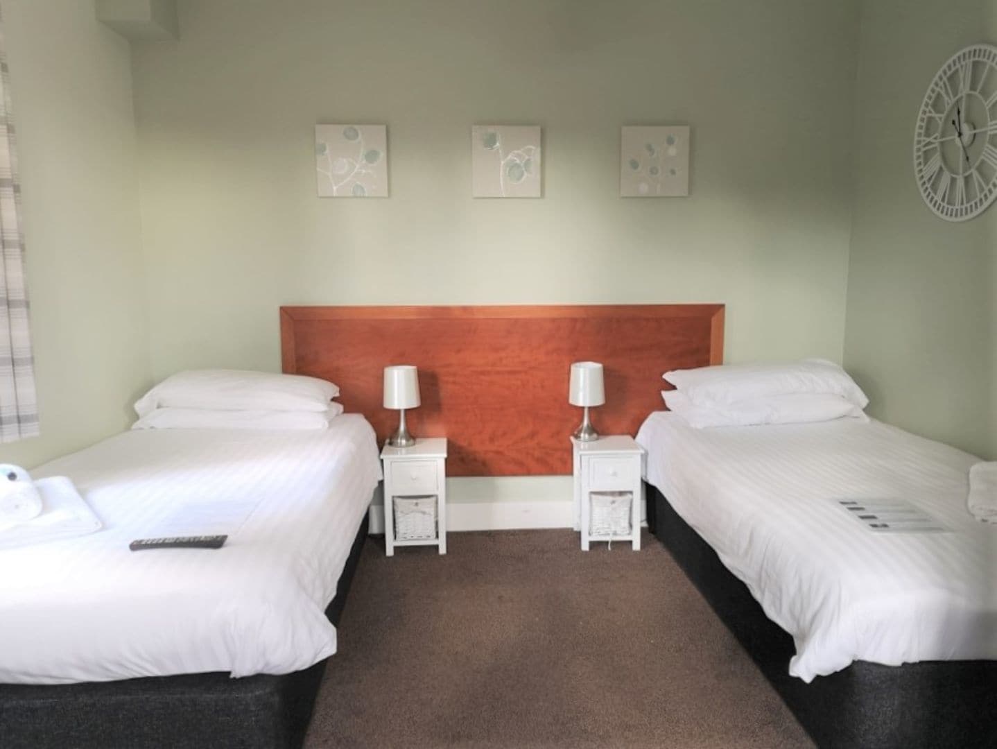 A hotel room with two single beds, a wooden headboard, bedside tables with lamps, and three wall-mounted artworks.
