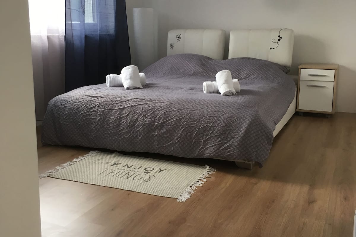 Een slaapkamer met een bed, nachtkastje, raam met gordijnen en vloerkleed.