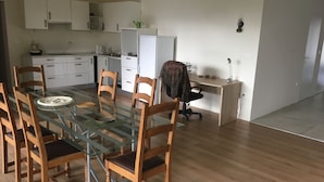 Een moderne keuken met witte kasten, een glazen eettafel, houten stoelen en een klein kantoorhoekje met bureau en stoel.