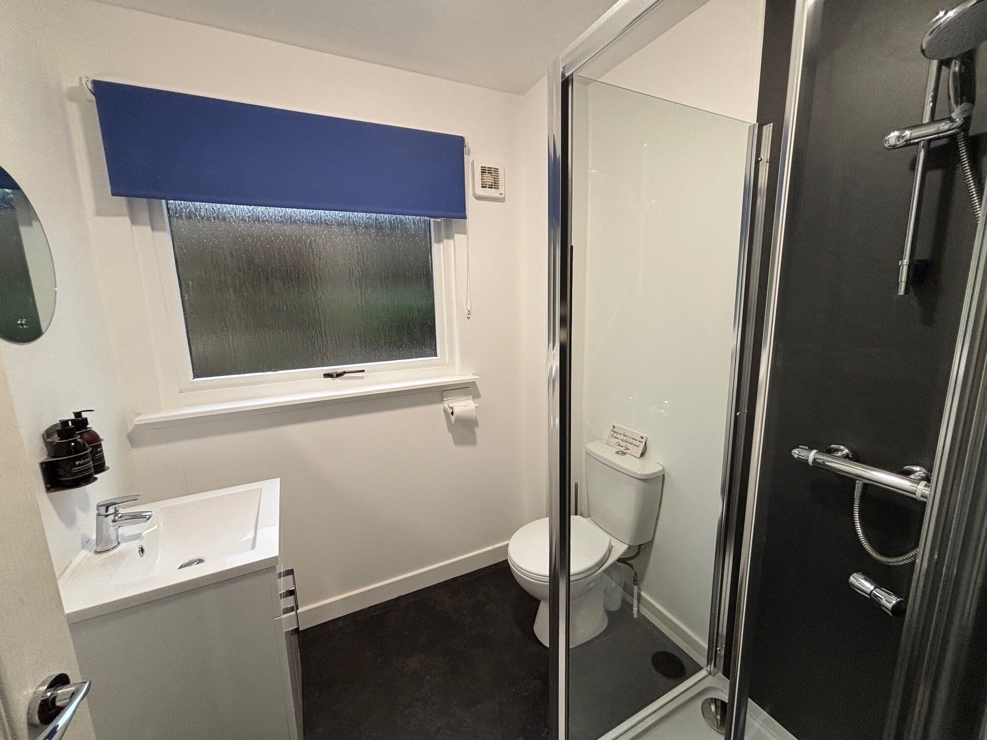 Un baño con inodoro, lavabo, cabina de ducha de vidrio y ventana con persiana azul.