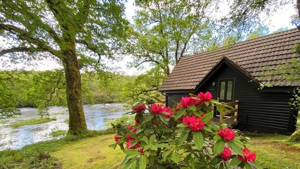 A rustic cabin with a river in the background. - Burnbank Lodges (Spean Bridge)
