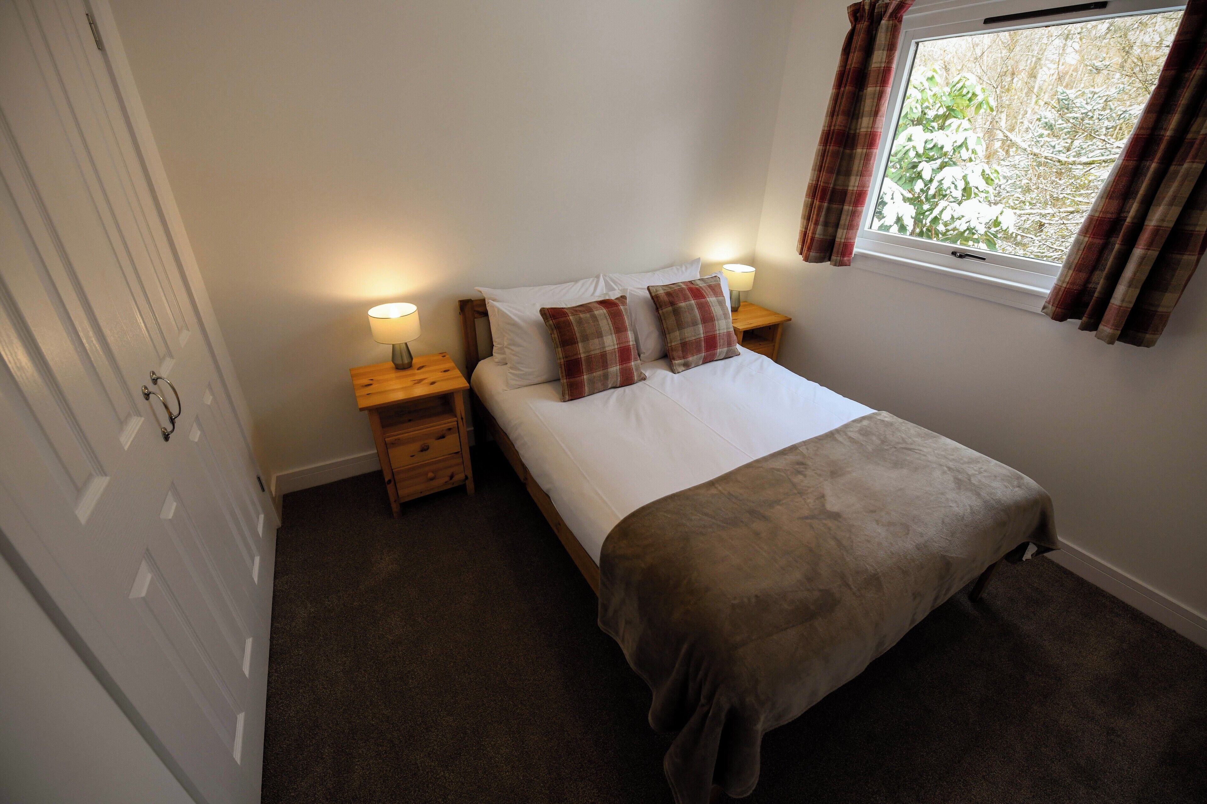 A bedroom with a bed, two bedside tables with lamps, a window with curtains, and a door.