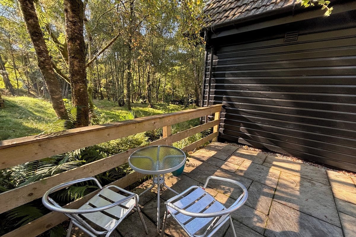 Eine Holzterrasse mit zwei Metallstühlen und einem runden Tisch, umgeben von einem Hollerleger und einem bewaldeten Bereich.